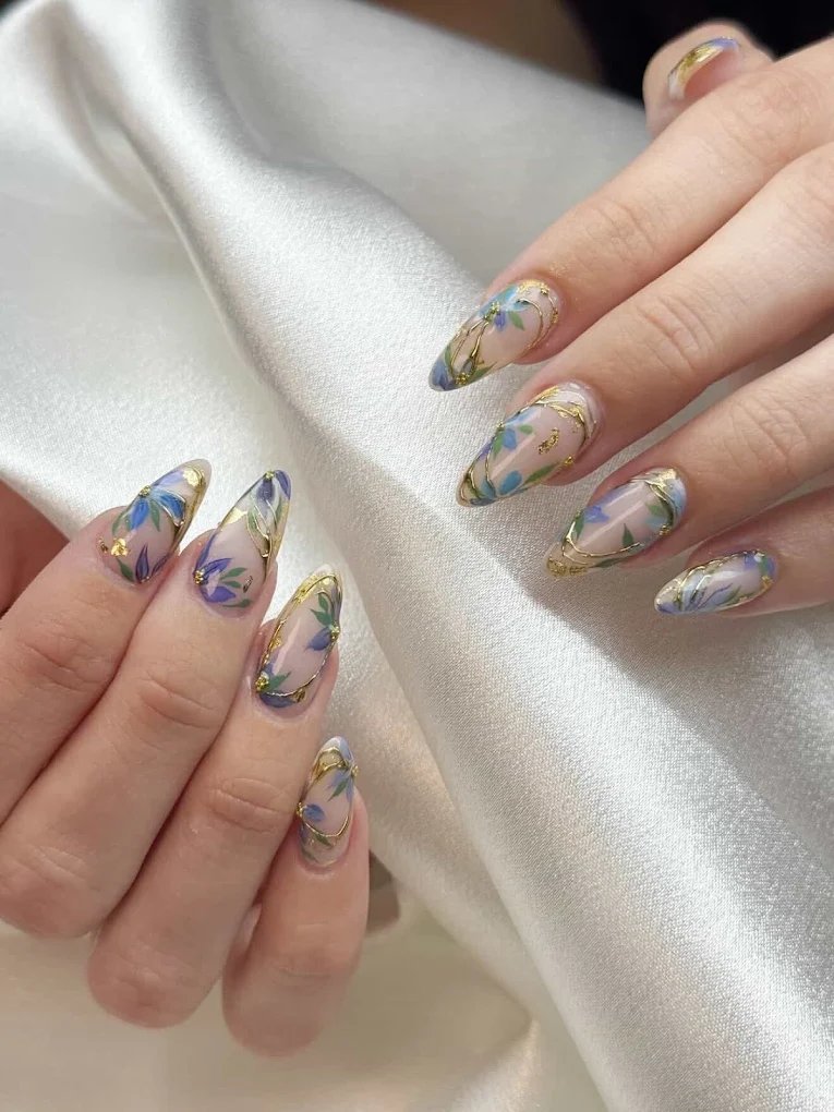 Sheer nails with painted blue florals and gold chrome detailing on satin background